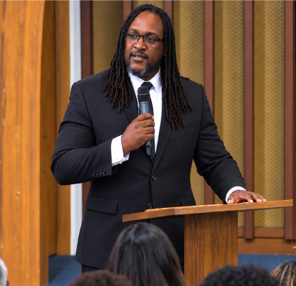 James Sanders speaks from a wooden podium in front of a group while wearing a black suit